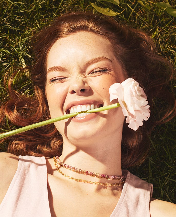 A woman lying on grass, playfully biting a flower stem, wearing a pink top with a beaded necklace, with the text 'SHOP CLEAN BEAUTY'